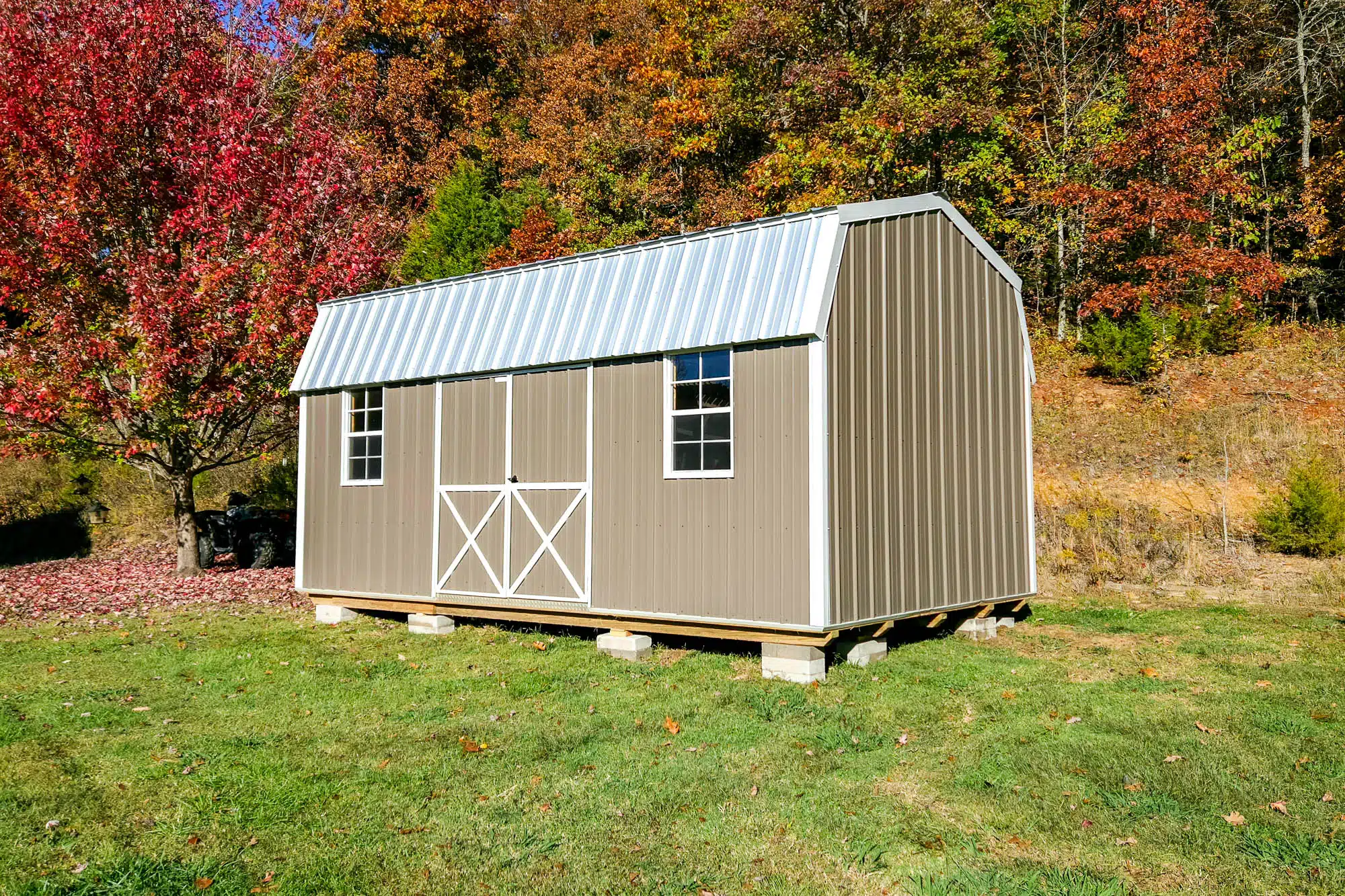 Tan lofted barn-style shed with white trim and metal roof, sitting on concrete blocks in a grassy yard with fall trees in the background.