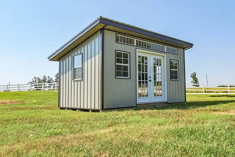 Modern portable shed with tan metal siding, transom windows, and double French doors, sitting on a grassy lawn with a white fence in the background.